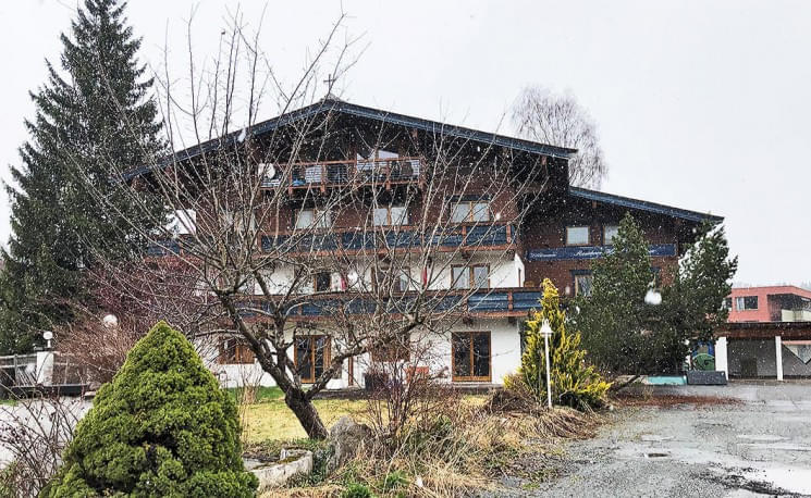 Ein dreistöckiges Holzgebäude im alpinen Stil mit verschneiten Bäumen im Vordergrund an einem grauen Tag.