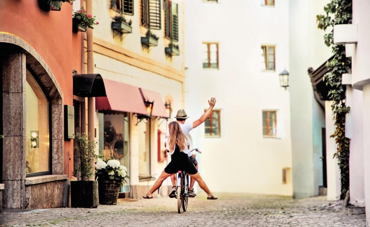 Ein Paar fährt fröhlich mit einem Fahrrad durch eine malerische Altstadt, umgeben von bunten Gebäuden und Kopfsteinpflaster. Eine friedliche, sonnige Szene.