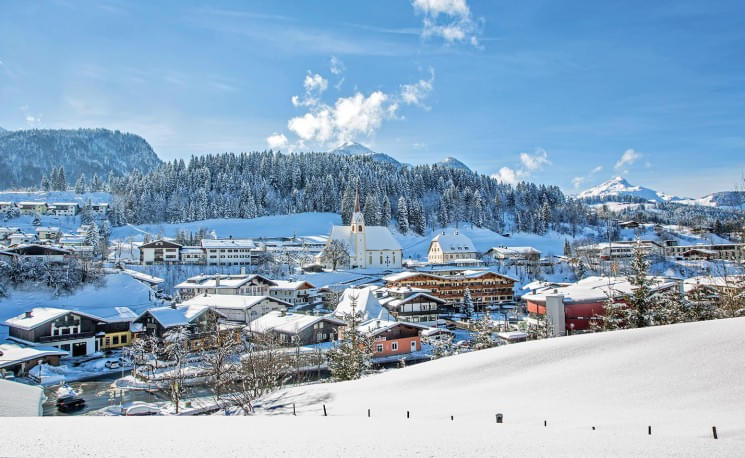 Eine malerische Winterlandschaft zeigt ein verschneites Dorf mit traditionellen Häusern, einer Kirche und bewaldeten Hügeln unter einem klaren blauen Himmel.