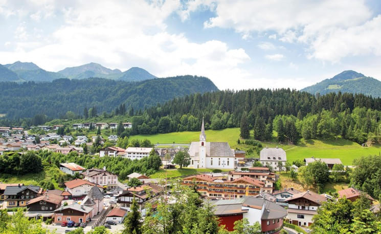 Panorama eines idyllischen Alpenortes mit Kirche im Zentrum, umgeben von grünen Wäldern und Bergen unter einem teilweise bewölkten Himmel.