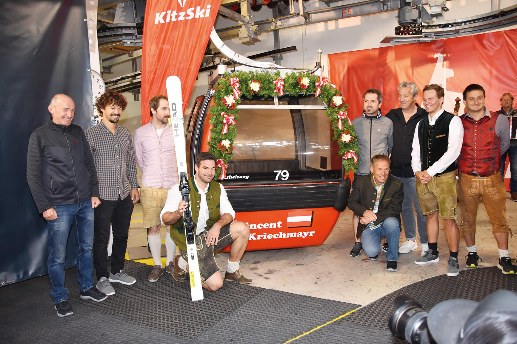 Eine Gruppe von Menschen steht vor einer geschmückten Skigondel mit Blumen. Einige tragen traditionelle Kleidung. Eine Person hält Ski in der Hand. Drinnen sind Kameras.