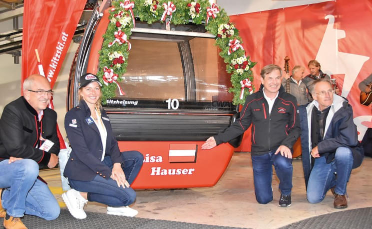 Eine Gruppe von vier Personen posiert vor einer rot-schwarzen Gondel, die festlich mit Blumen geschmückt ist. "Lisa Hauser" ist auf der Gondel vermerkt.