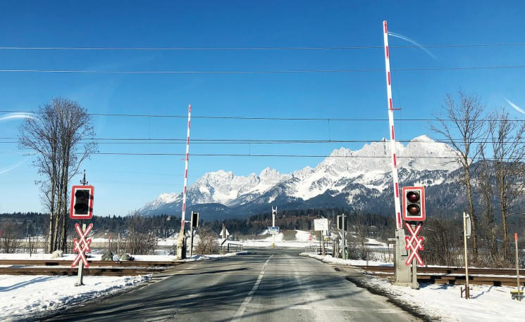 Schneebedeckte Bahnübergang in Alpenkulisse mit Lichter-Signalen und klarblauem Himmel im Hintergrund. Ein ruhiger, winterlicher Tag.