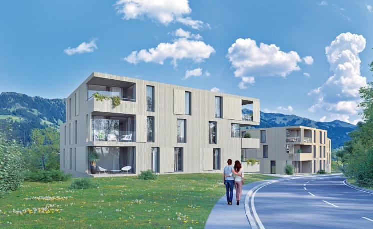 Moderne, kubische Wohngebäude mit großen Fenstern in einer grünen Landschaft. Zwei Personen gehen auf einem Gehweg, blauer Himmel mit weißen Wolken im Hintergrund.