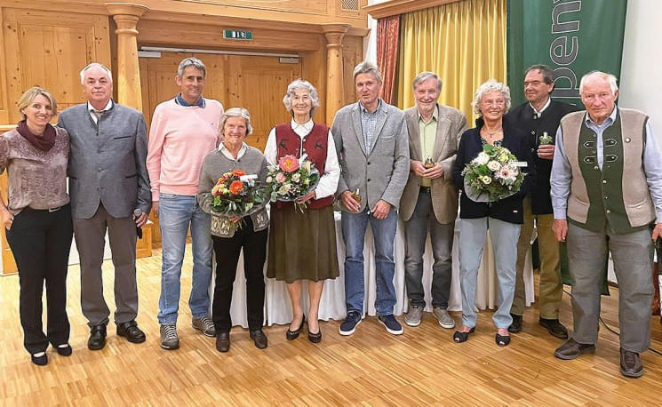 Eine Gruppe von älteren Personen steht lächelnd in einem Saal, einige halten Blumensträuße. Im Hintergrund sind Holzverkleidung und Vorhänge zu sehen.