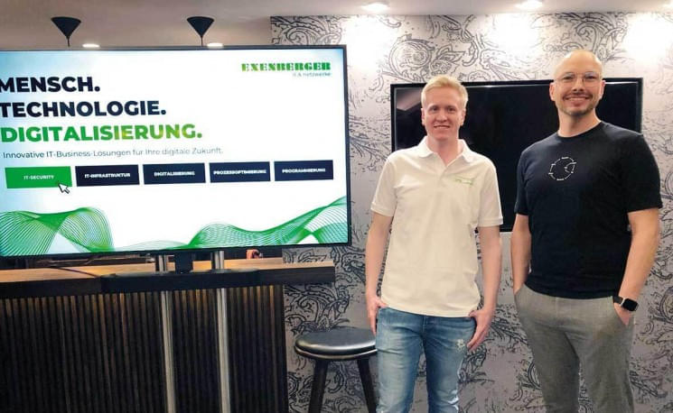 Zwei Menschen stehen lächelnd vor einem Bildschirm, auf dem "Mensch. Technologie. Digitalisierung." zu lesen ist. Die Umgebung ist modern gestaltet.