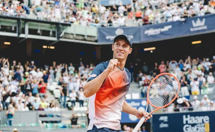 Ein Tennisspieler jubelt auf dem Spielfeld und hält einen Schläger, während das Publikum im Stadion applaudiert. Er trägt Sportkleidung und eine Kappe.