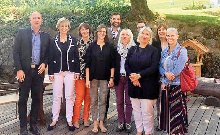 Eine Gruppe von zehn Erwachsenen steht auf einer Holzterrasse im Freien. Sie sind lächelnd und in Freizeit- oder Businesskleidung gekleidet.