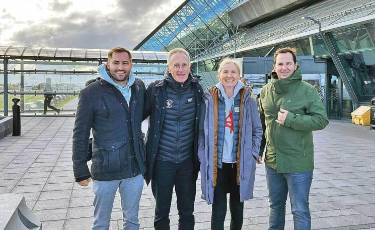 Vier Personen stehen in Outdoor-Kleidung auf einem Flughafengelände vor einem modernen Gebäude mit Glasfassade und lächeln in die Kamera.