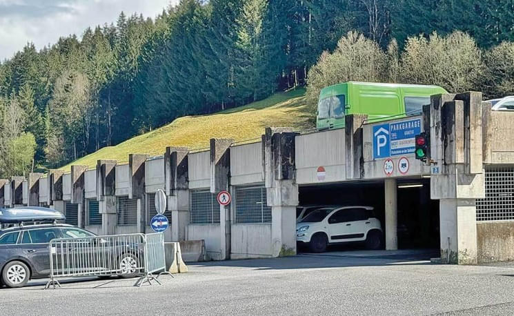 Ein Parkhaus am Waldrand mit einem grünen LKW oben. Ein weißer Van fährt heraus, während ein weiteres Auto am Eingang parkt. Verkehrszeichen sind sichtbar.