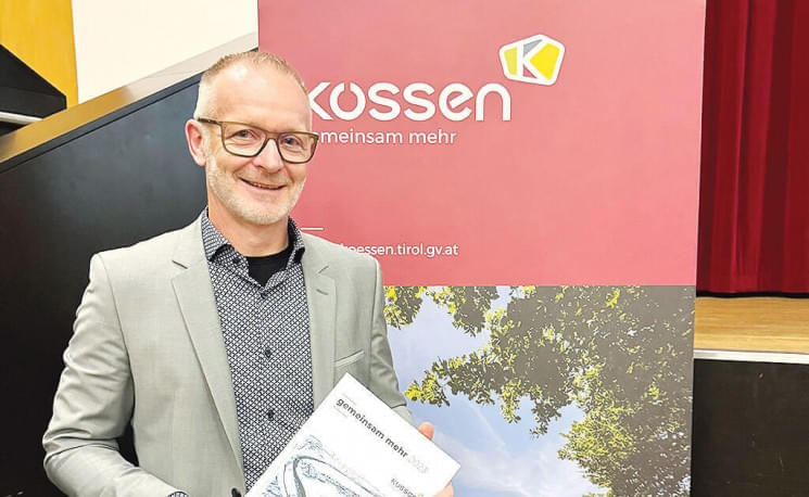 Ein Mann mit Brille und grauem Anzug steht vor einem Kossen-Banner und hält ein Buch in den Händen.