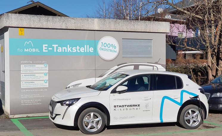Zwei weiße Elektroautos parken vor einer E-Tankstelle mit der Aufschrift "100% Ökostrom" und dem Logo der Stadtwerke. Ein sonniger Tag.