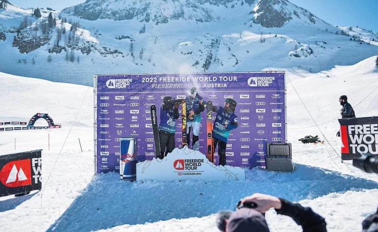 Drei Personen stehen auf einem Podium vor einer Bergkulisse auf einem Skievent, umgeben von Schnee und Sponsorenwerbung. Es handelt sich um eine Siegerehrung.