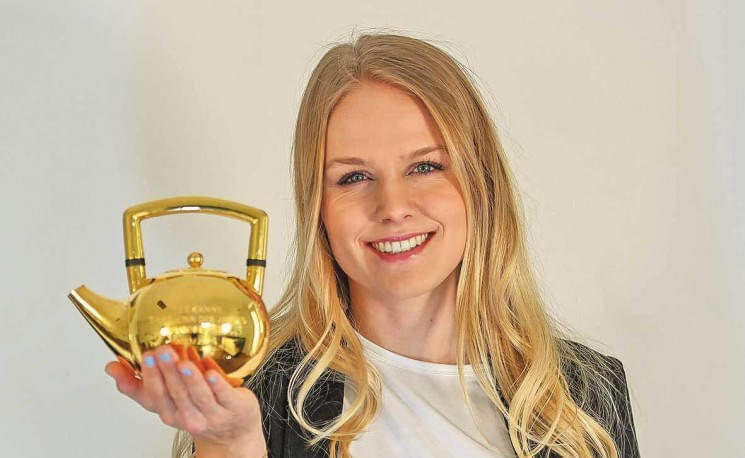 Frau mit langen blonden Haaren hält eine glänzende goldene Teekanne und lächelt in die Kamera. Sie trägt eine schwarze Jacke über einem weißen Oberteil.