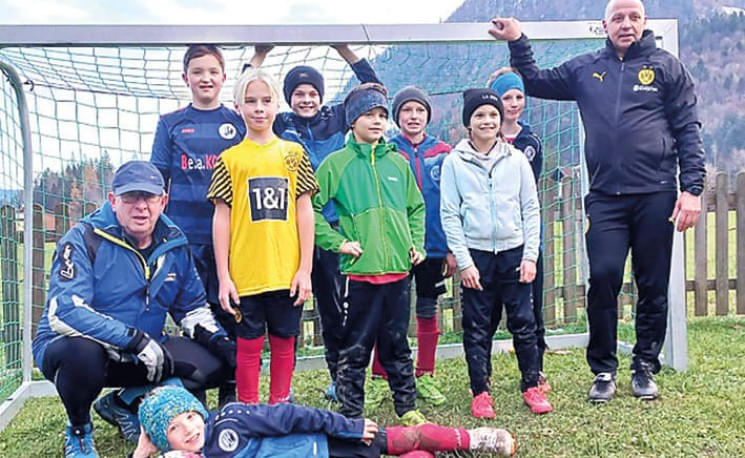 Eine Gruppe von jungen Fußballspielern und zwei Trainern posiert fröhlich vor einem Fußballtor auf einem Feld mit Bergen im Hintergrund.
