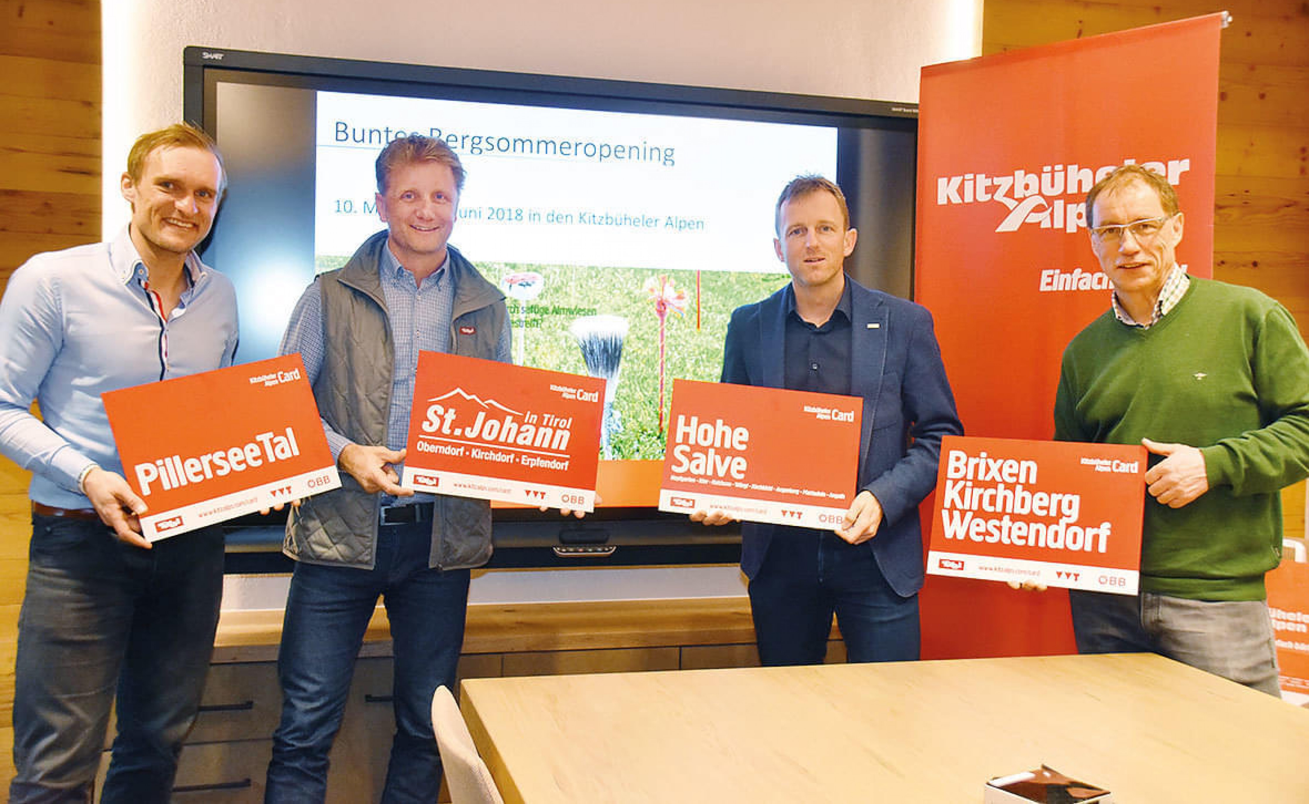 Vier Männer präsentieren Werbeschilder für Regionen in den Kitzbüheler Alpen vor einem Bildschirm in einem Konferenzraum.