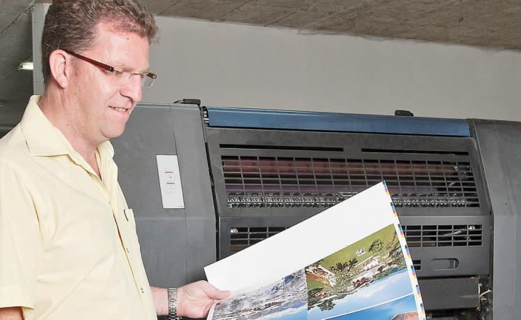 Ein Mann steht vor einem großen Drucker und betrachtet ein Blatt mit bunten Bildern. Er trägt ein helles Hemd und eine Brille, während er den Druck prüft.