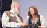 Ein Mann mit Bart und eine Frau in traditioneller Kleidung unterhalten sich lächelnd auf einer Bühne oder bei einer Veranstaltung.