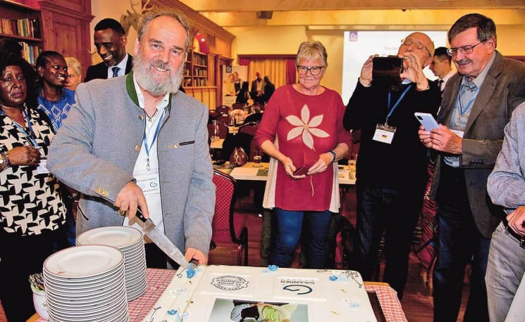 Eine Gruppe lachender Menschen steht um eine festliche Torte, während ein Mann in einem grauen Sakko sie anschneidet. Sie befinden sich in einem gemütlichen Raum.