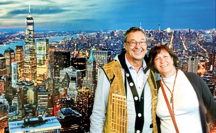 Ein lächelndes Paar steht vor einem nächtlichen Stadtpanorama mit beleuchteten Wolkenkratzern, darunter ein hoch aufragendes Gebäude im Vordergrund.