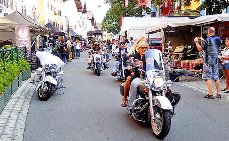 Eine Gruppe von Motorradfahrern fährt durch eine belebte Straße mit Marktständen und Zuschauern daneben bei sonnigem Wetter.
