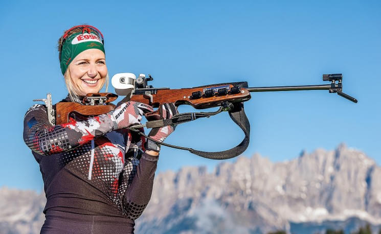 Eine Biathletin zielt mit einem Gewehr auf eine Zielscheibe. Sie trägt einen gemusterten Rennanzug und Stirnband, im Hintergrund sind schneebedeckte Berge sichtbar.
