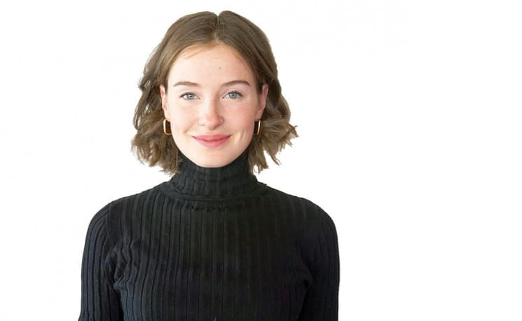 Junge Frau mit schulterlangem braunen Haar trägt einen schwarzen Rollkragenpullover und lächelt vor einem weißen Hintergrund in die Kamera.