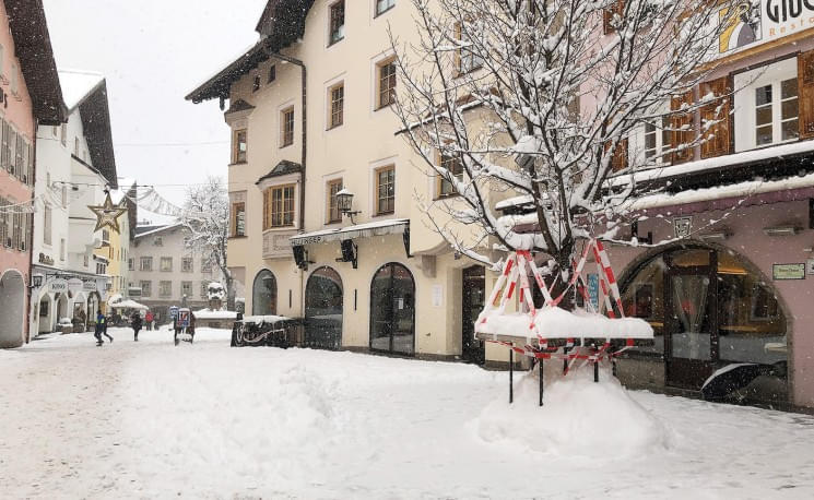 Verschneite Fußgängerzone in einer malerischen Stadt, umgeben von Alpenhäusern und einem geschmückten Baum. Menschen spazieren entlang der Straße.