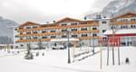 Ein großes, mehrstöckiges Gebäude im alpinen Stil mit Holzbalkonen, umgeben von Schnee und Bergen im Hintergrund. Es wirkt wie ein Hotel oder Ferienresort.
