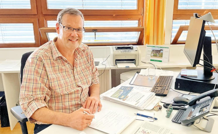 Ein Mann sitzt an einem Schreibtisch, lächelt in die Kamera und arbeitet an Dokumenten. Im Hintergrund sind ein Drucker und ein Monitor zu sehen.