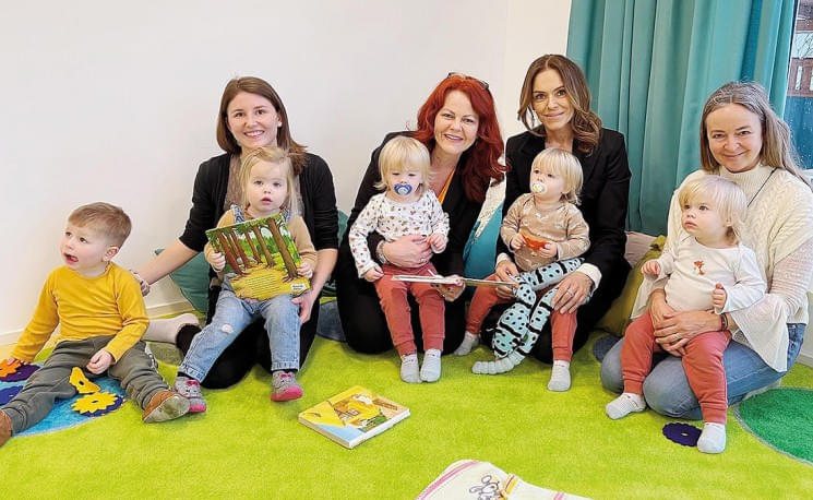 Erwachsene Frauen sitzen mit kleinen Kindern auf einem bunten Teppich, während sie Bücher halten. Im Hintergrund hängt ein türkisfarbener Vorhang.