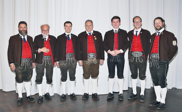 Sieben Männer in traditionellen bayrischen Trachten stehen nebeneinander vor einem weißen Hintergrund und lächeln. Sie tragen Lederhosen, rote Westen und dunkle Jacken.