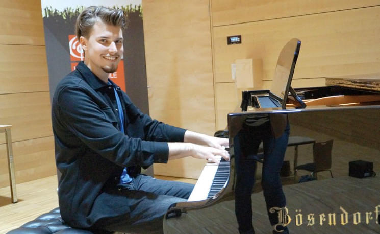 Junger Mann in schwarzem Hemd spielt Klavier in einem modernen Raum mit Holzverkleidung. Im Hintergrund ist ein Musiklogo zu sehen.