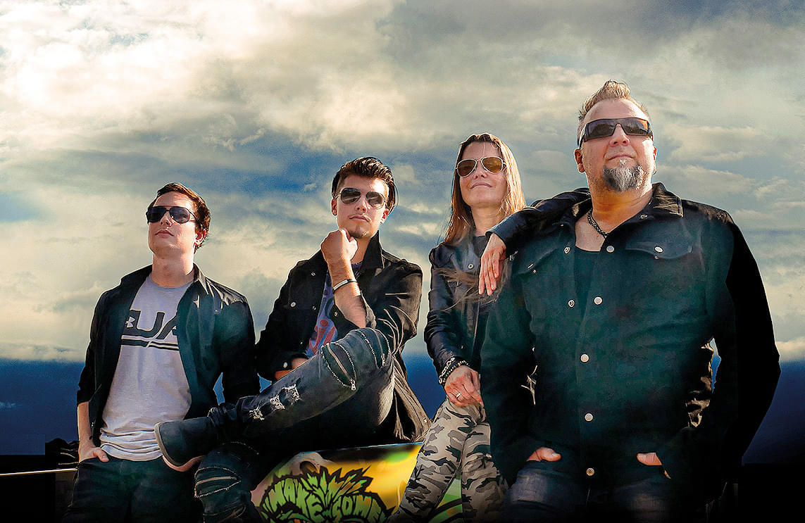 Vier Personen posieren lässig unter einem bewölkten Himmel. Alle tragen Sonnenbrillen und dunkle Kleidung. Die Stimmung ist entspannt und selbstbewusst.