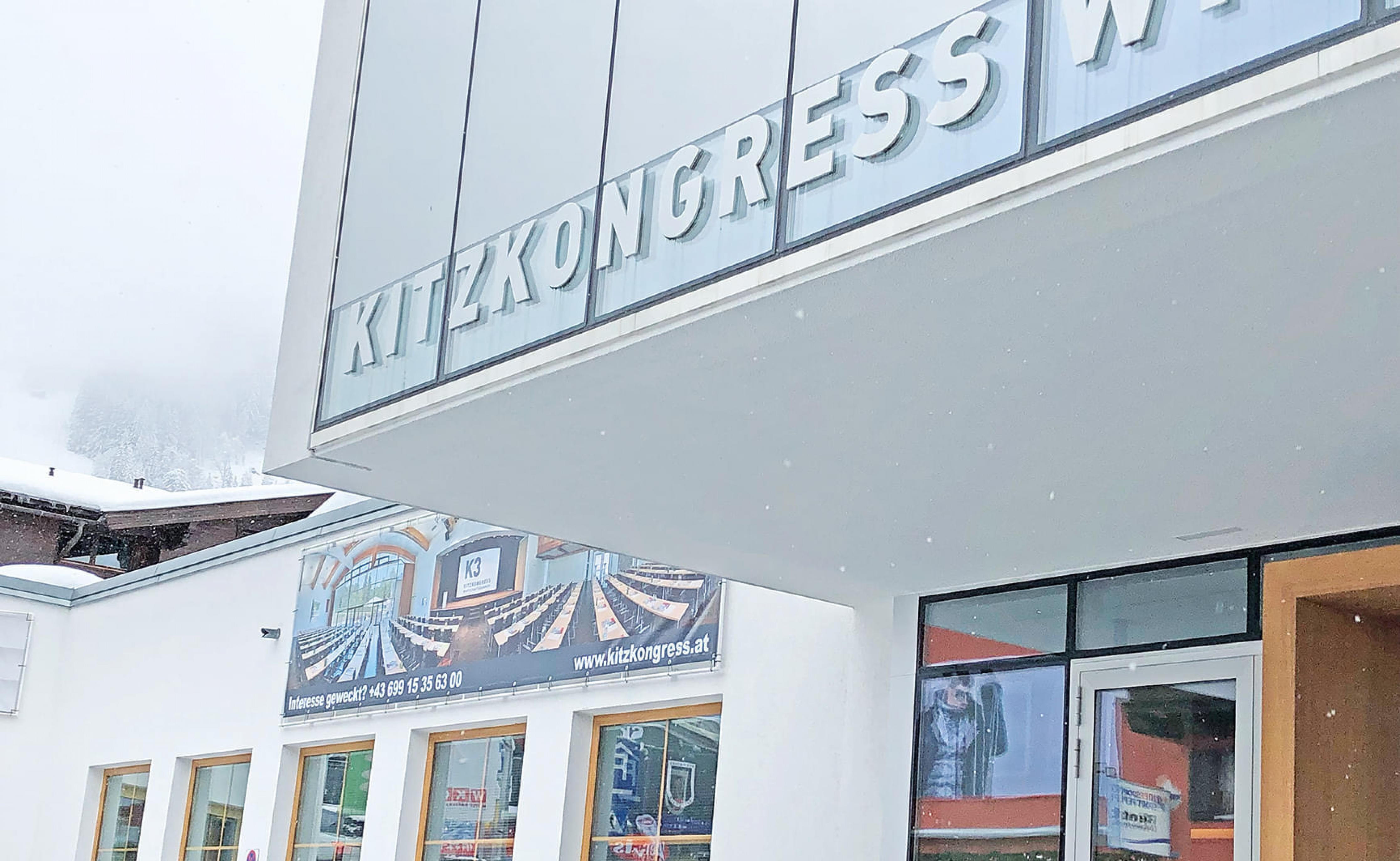 Eingang eines Kongresszentrums mit der Aufschrift „Kitzkongress“; moderne Glasfassade und Beschilderung, schneebedeckte Landschaft im Hintergrund.