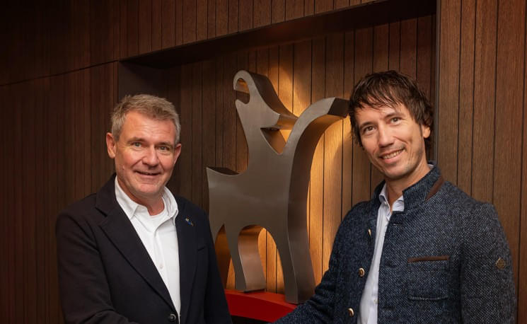 Zwei Männer in Anzügen schütteln sich die Hände und lächeln in die Kamera. Sie stehen vor einer modernen Skulptur in einem Holzvertäfelten Raum.