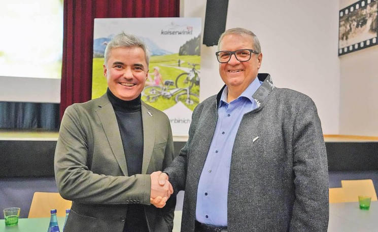 Zwei Männer in Anzügen schütteln sich die Hand in einem Saal, lächeln und stehen vor einem Plakat mit Outdoor-Szenen. Ein roter Vorhang ist im Hintergrund.