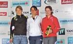 Drei Personen posieren vor einer Sponsorenwand. Zwei tragen Sportbekleidung, eine hält eine Trophäe. Sie lächeln und scheinen an einem Sportevent teilzunehmen.