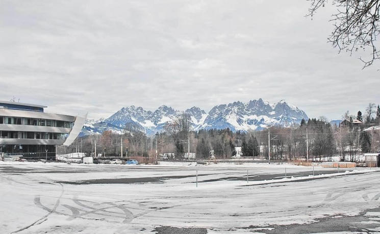 Winterliche Landschaft mit schneebedeckten Bergen im Hintergrund und einem modernen Gebäude links im Bild. Der Himmel ist bewölkt und der Boden ist verschneit.