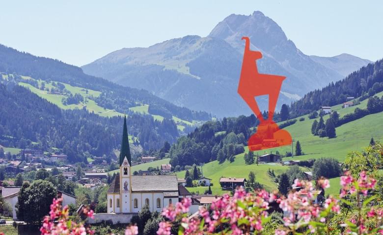 Landschaft mit grünem Tal, Kirche im Vordergrund, blühenden Blumen und bergiger Hintergrund. Eine stilisierte, orangefarbene Figur steht zentral im Bild.