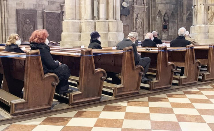 Menschen sitzen mit Abstand in einer Kirchenbank, einige tragen Hüte. Die Kirche hat steinerne Säulen und verzierte Wände im Hintergrund.