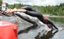 Sportler in einem schwarzen Neoprenanzug springt kopfüber in einen See, umgeben von Bäumen. Ein bunter Bojenmarker ist im Vordergrund zu sehen.