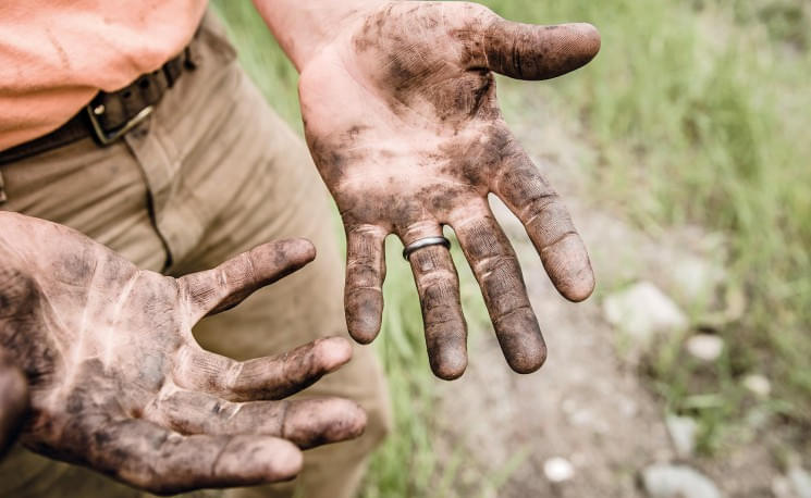 Schmutzige Hände eines Menschen mit Ehering, im Freien aufgenommen. Der Hintergrund zeigt unscharfes Grün und Erde. Die Person trägt beige Hose.