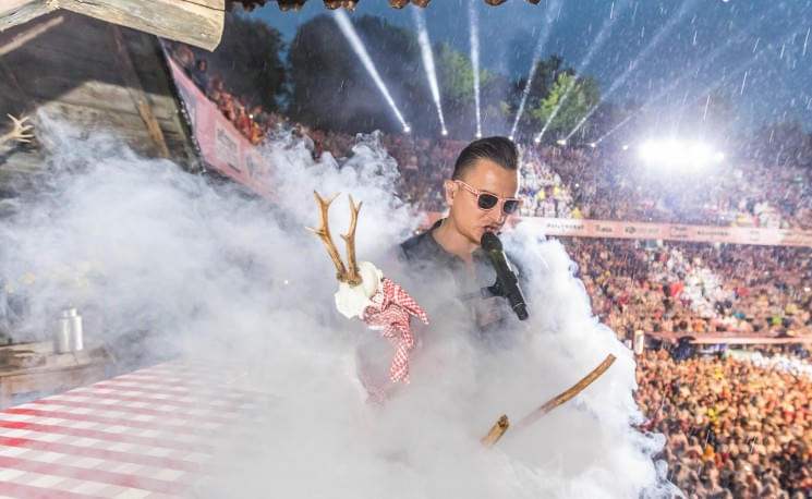 Ein Sänger mit Sonnenbrille performt auf einer Bühne im Freien, umgeben von Nebel und Lichteffekten, während ein großes Publikum zusieht.