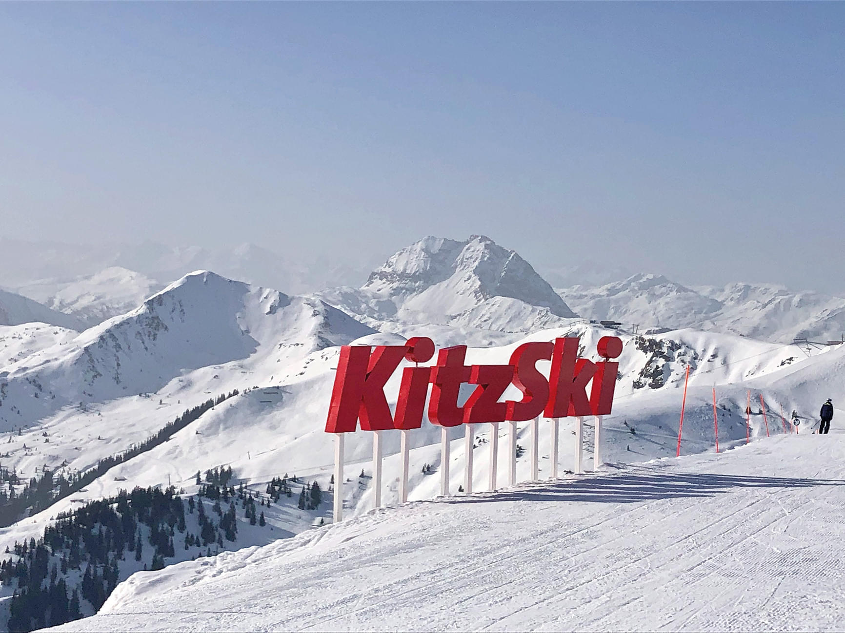 Verschneite Berge bei klarem Himmel, im Vordergrund steht ein großes Schild mit der Aufschrift "KitzSki". Die Landschaft wirkt idyllisch und einladend.
