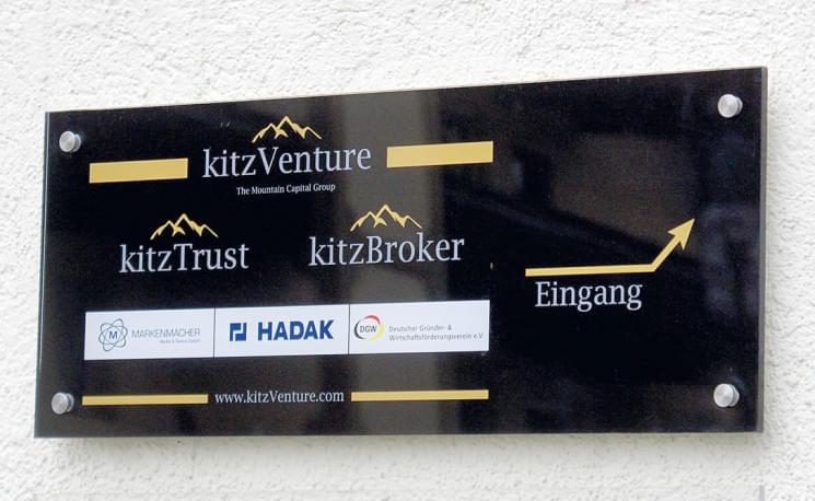 Ein Schild an einer Wand zeigt verschiedene Firmenlogos wie kitzVenture, kitzTrust und kitzBroker mit einem Pfeil und dem Wort "Eingang".