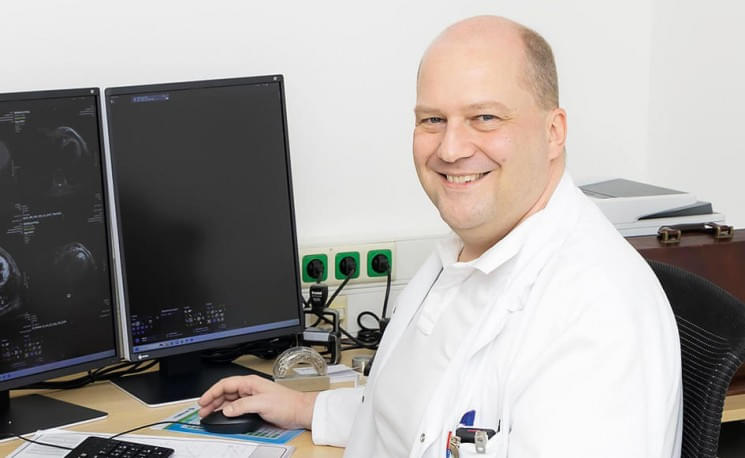 Ein Mann in einem weißen Kittel sitzt lächelnd an einem Schreibtisch vor zwei Monitoren in einem Büro oder Laborumgebung.