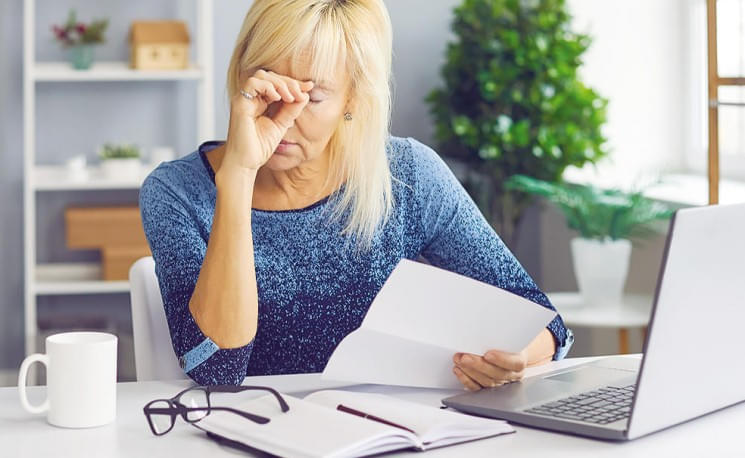 Eine Frau sitzt an einem Schreibtisch vor einem Laptop. Sie reibt sich die Augen und hält einen Brief in der Hand. Brille und Notizbuch liegen daneben.