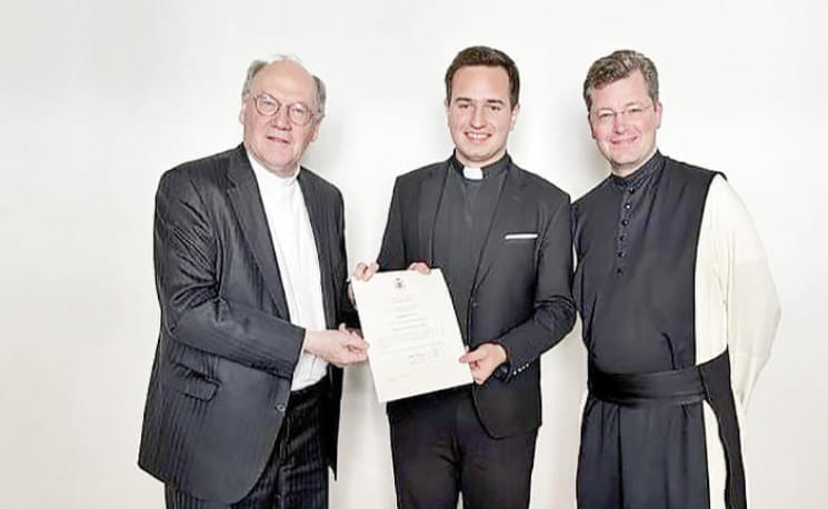 Drei Männer in kirchlicher Kleidung halten ein Dokument vor einem weißen Hintergrund. Der mittlere Mann trägt einen Anzug mit Kollar.