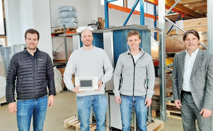 Vier Männer stehen in einem Lagerraum, zwei von ihnen halten ein Gerät. Sie tragen legere Kleidung und lächeln in die Kamera.